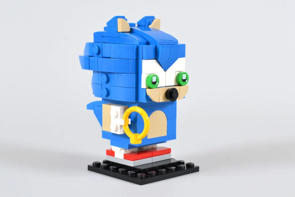 A LEGO BrickHeadz model of Sonic the Hedgehog, featuring Sonic’s iconic blue spiky hair, green eyes, and red shoes, holding a yellow ring and standing on a black baseplate.