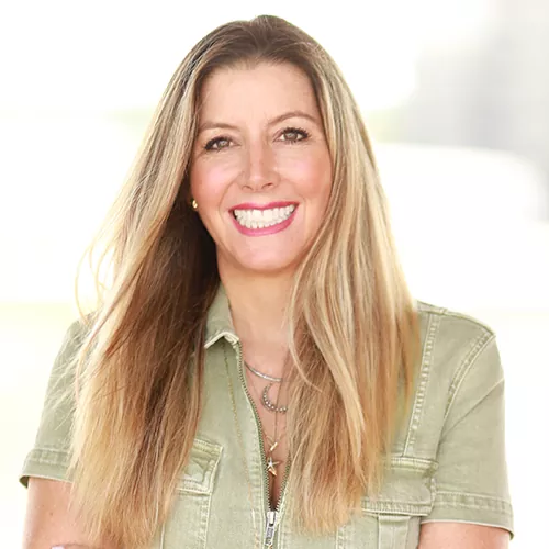 A smiling woman with long blonde hair, wearing a light green casual jacket and layered necklaces, exuding warmth and confidence in a bright, outdoor setting.
