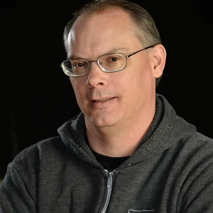 A casual portrait of Tim Sweeney, wearing glasses and a dark hoodie, with a neutral expression against a black background.